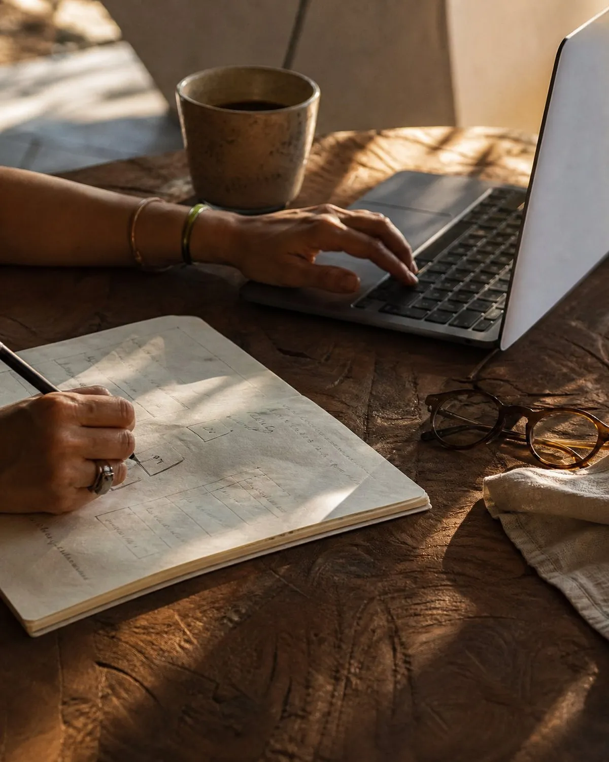 Hands at a carved teak table, one on a laptop trackpad and one sketching a diagram in a notebook