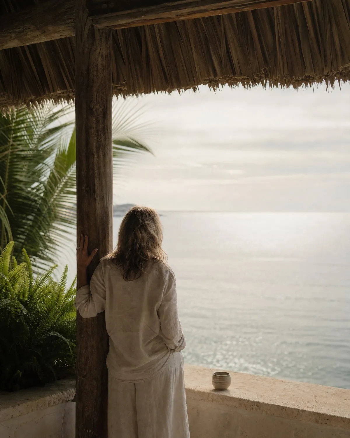 A woman in linen standing at the open edge of a palapa-roofed room, looking out at calm water in early morning light